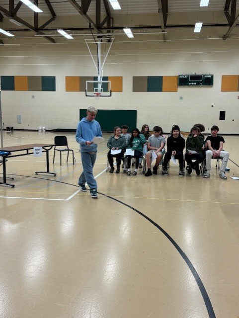 An individual stands in a gymnasium with a basketball hoop, addressing a group of seated students.
