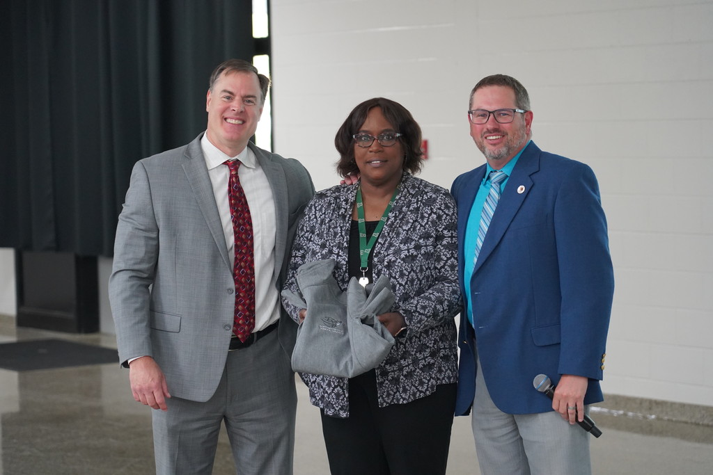 Three people stand in front of a white wall, each dressed in professional attire. The woman holds a jacket.