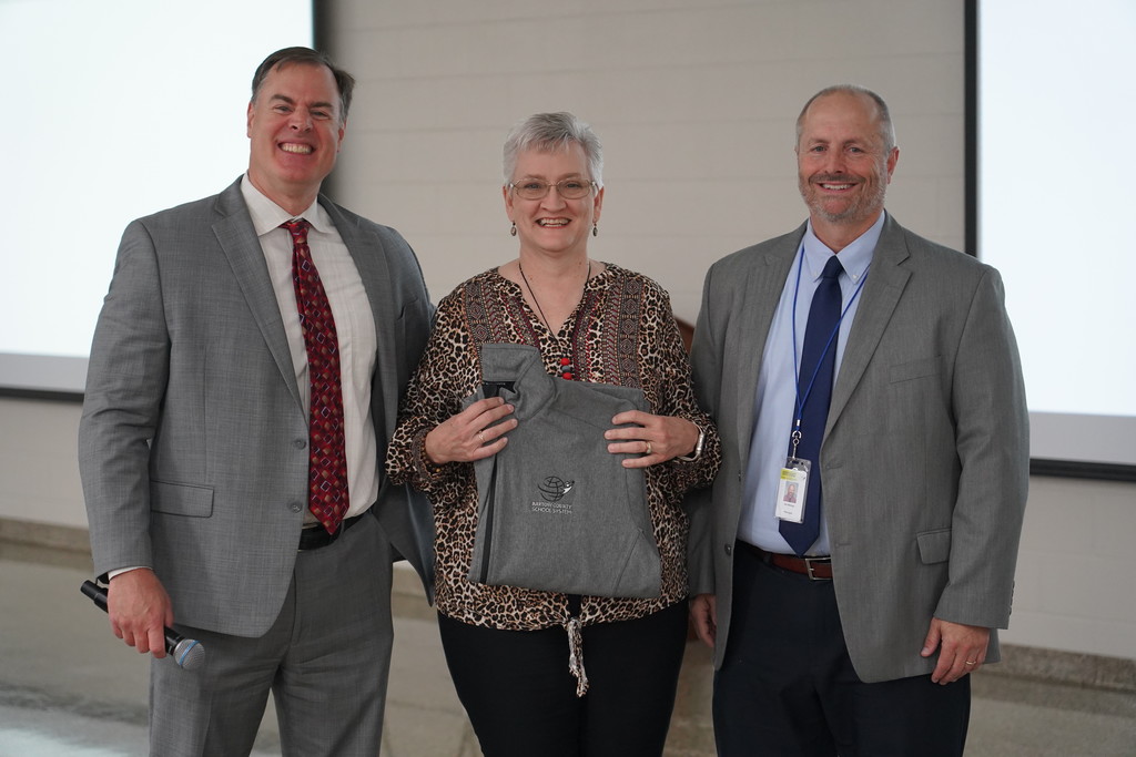 Three individuals in suits stand close together; the central person holds a gray garment. One wears a blue tie, another a red tie, and the central person wears glasses.