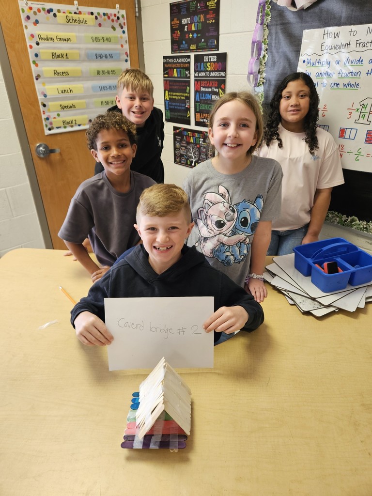 Five children in a classroom setting, holding a paper sign and standing around a table.