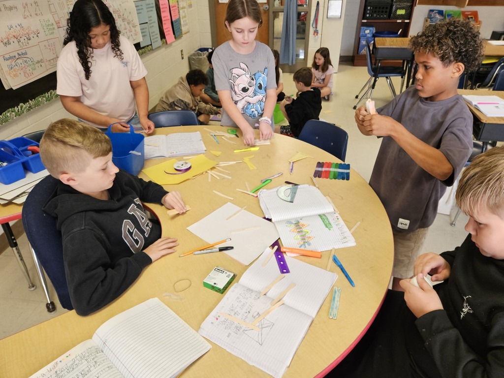 Several children at a round table in a classroom work on a project. One child holds a marker.