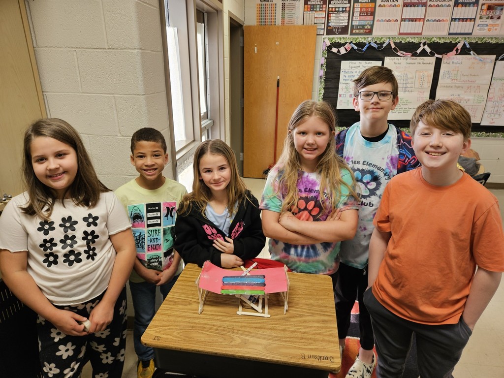 Six children, including girls and boys, stand around a small wooden table with a model on it in a classroom.