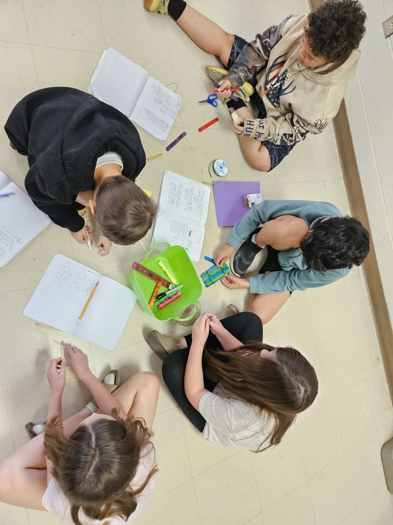 Five young people sit on the floor in a circle, engaged in a group project. Papers and markers are scattered around them.