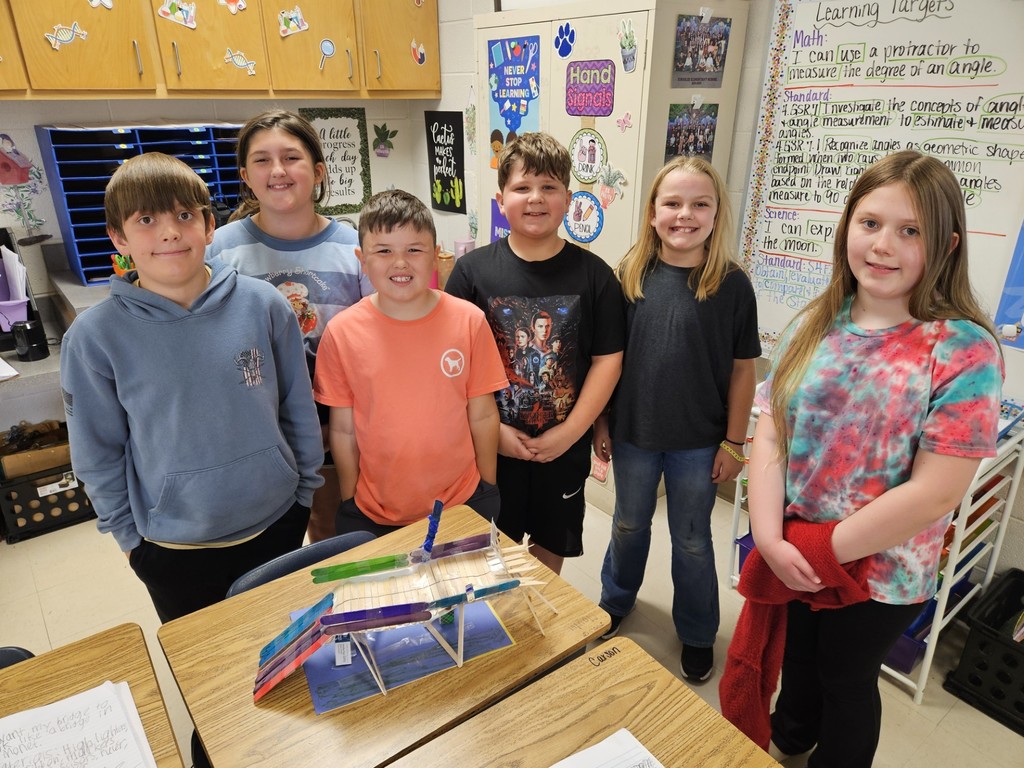 A group of children stands in a classroom around a table displaying a model. They appear to be students.