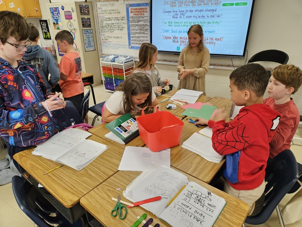 A classroom with students seated at desks. A teacher stands in front of a screen. Papers and scissors are on the desks.