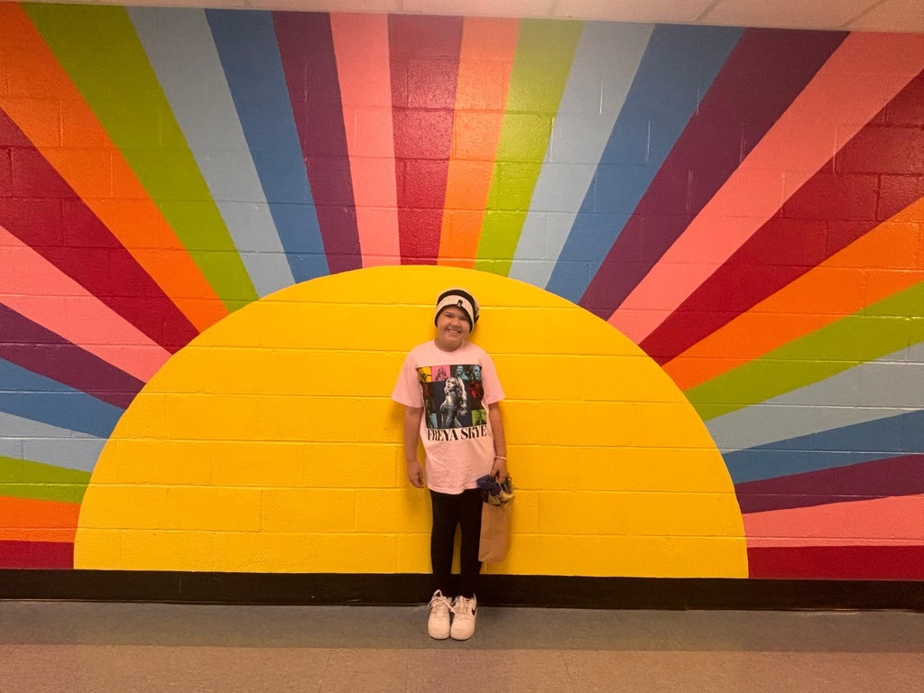 A girl  in a pink shirt stands in front of a wall with a colorful sunburst design.