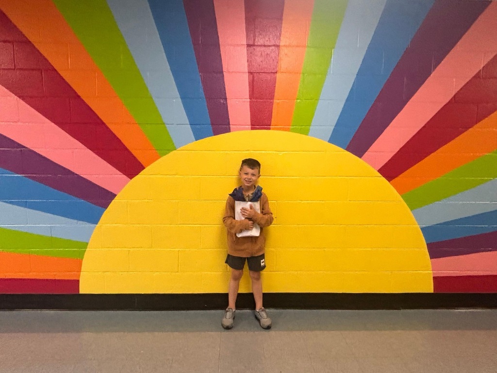 A young boy with a book stands in front of a colorful sun mural on a wall.