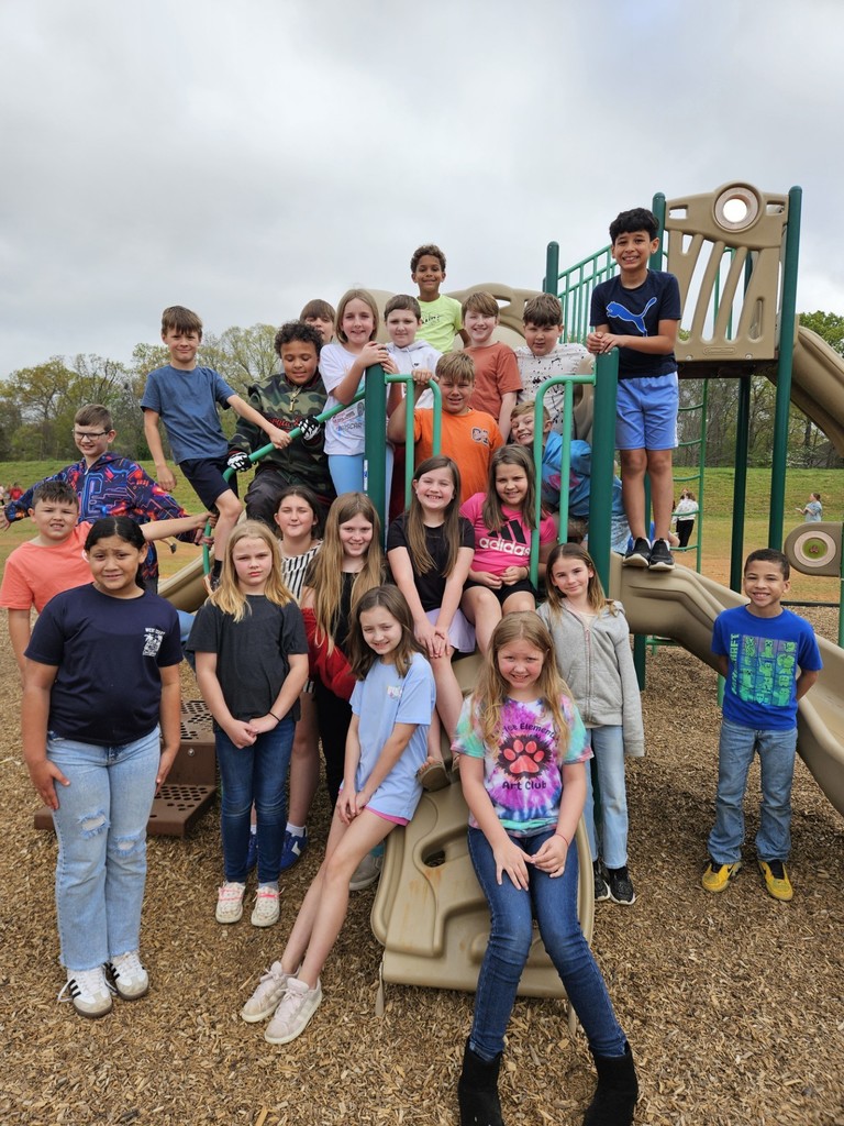 Several children pose for a picture in an outdoor playground with wooden structures and slides under a cloudy sky.