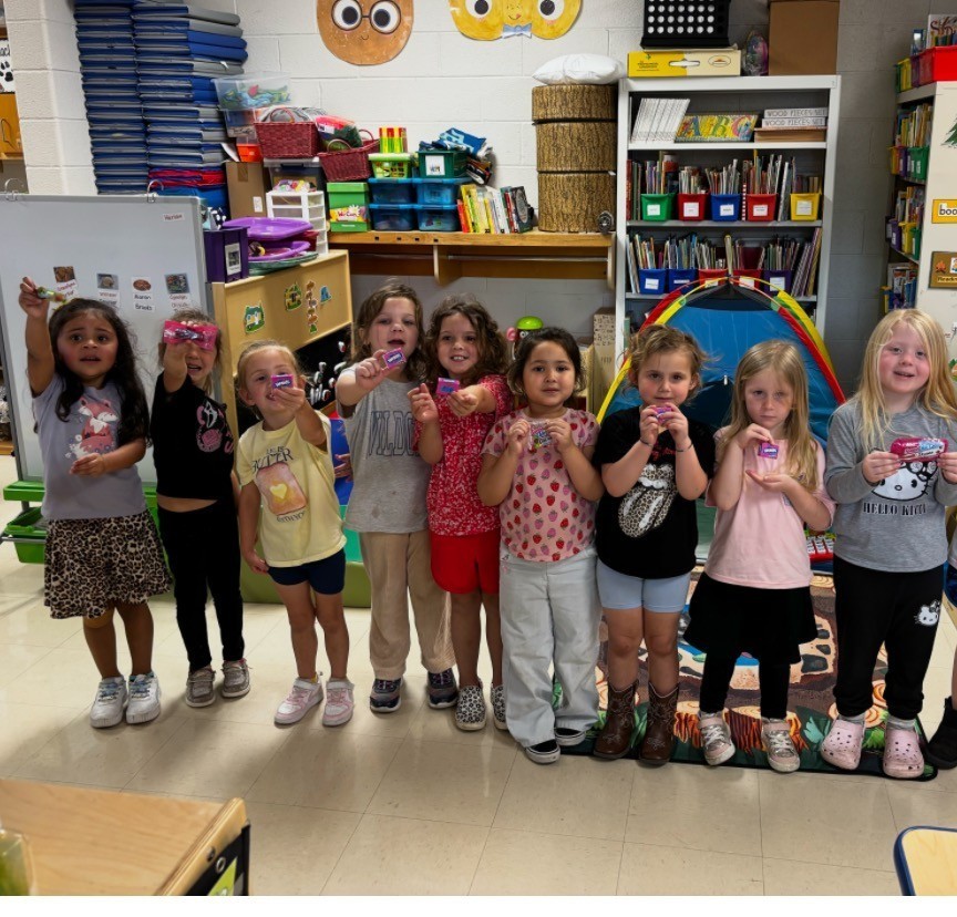 Group of children, possibly students, standing in a classroom with various items on shelves and a rug on the floor.