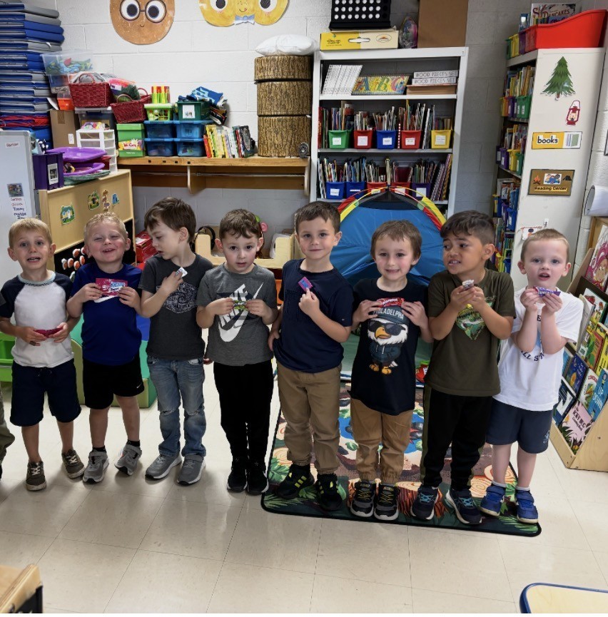 A group of children in a classroom, holding a small object. The room is filled with bookshelves and shelves.