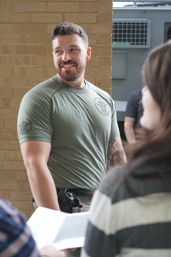 A man in a green shirt stands near a brick wall, smiling and looking at a woman in a striped shirt.