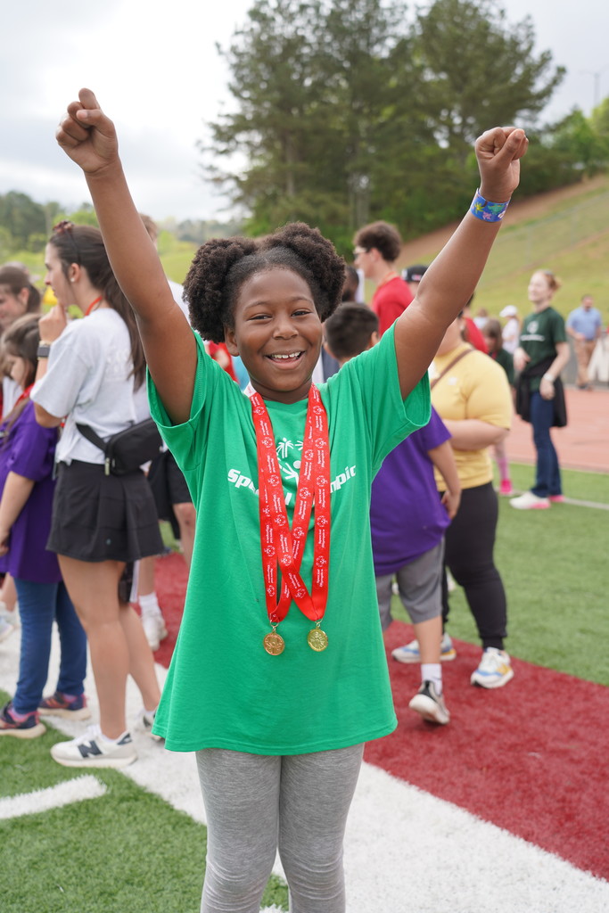 A girl with her arms raised, wearing a green shirt and medals, stands on a football field with other people.