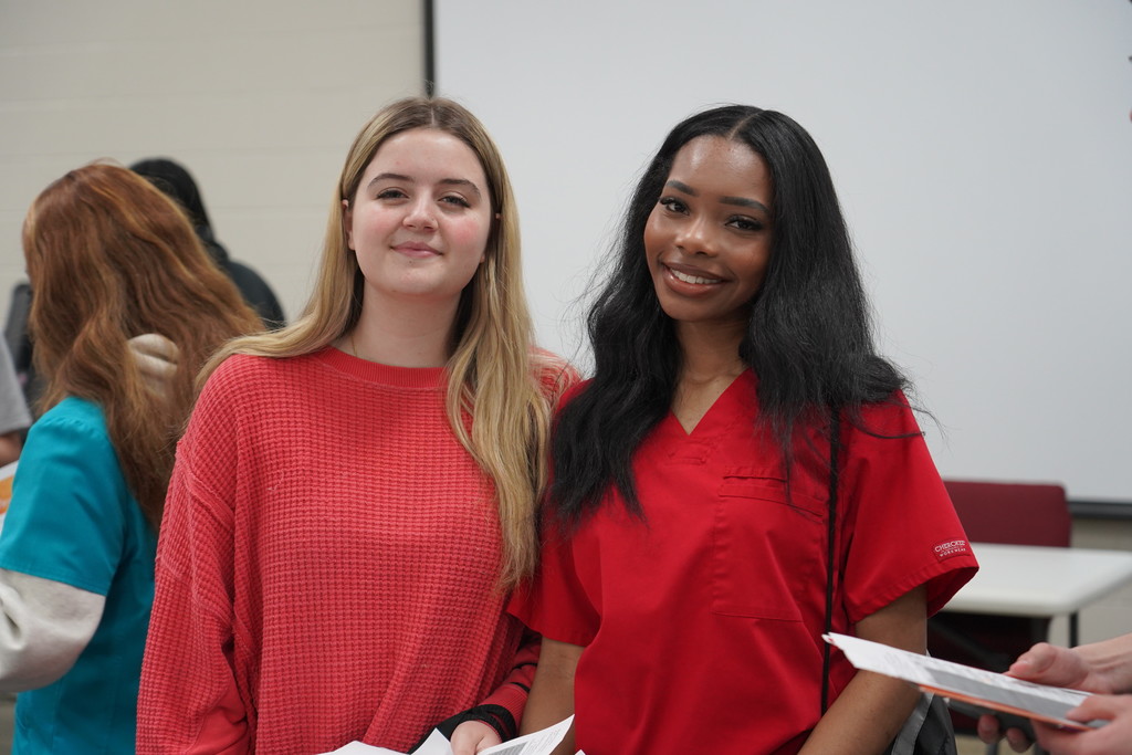 Two women in red shirts stand together in a classroom. One holds a paper. Behind them, a white board and chairs are visible.