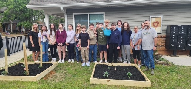 A group of people, including children, stand in front of a house. They are posing in front of three wooden garden beds.