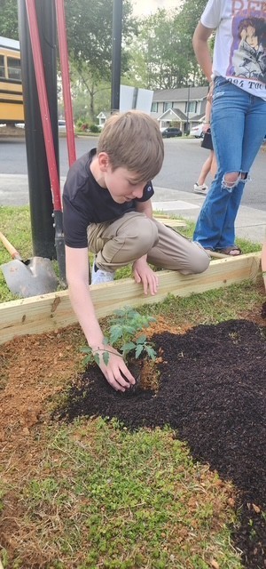 A young boy kneels in a garden bed, planting a small plant. A shovel is nearby. A person stands behind him.