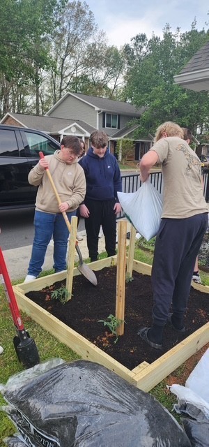 A group of people work in a raised garden bed, one holding a shovel, another holding a plant.