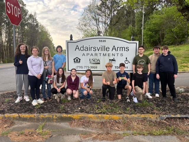 A group of individuals stands in front of a white sign with the words "Adairsville Arms Apartments".