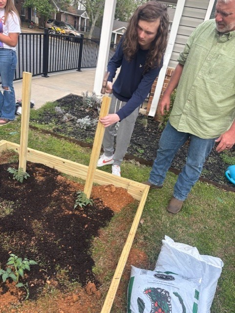 Two people working in a raised garden bed; one holding a stick and another holding a bag of soil.