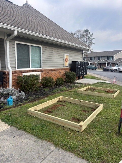 Two wooden garden boxes with soil in a yard in front of a house. There is a bush and a mailbox.