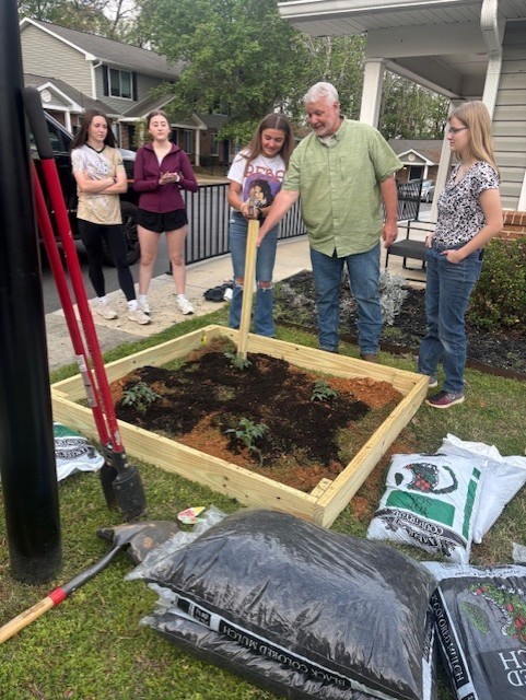A man with a shovel stands near a wooden raised bed filled with soil. Four women are around him.