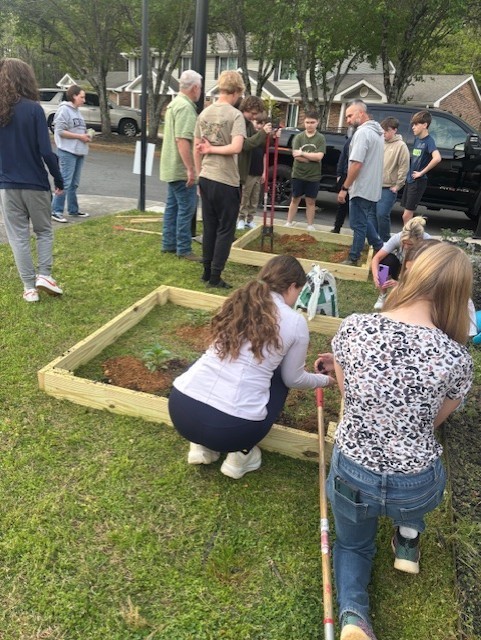 Several people work in raised garden plots, some kneeling, others standing; a car is parked in the background.