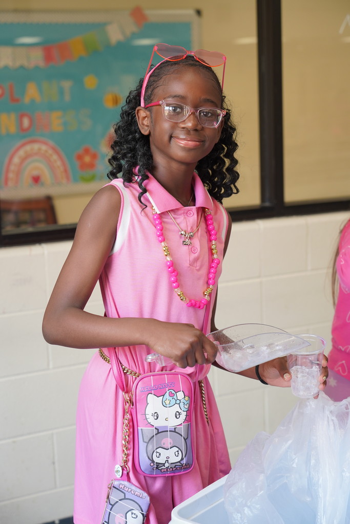 A girl in pink holds plastic, standing near a white wall with a banner. She wears a pink dress and has glasses and a necklace.