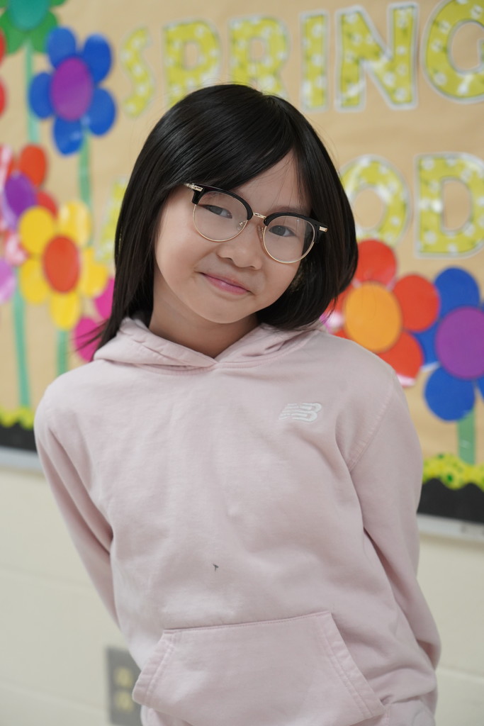 Girl with black hair wearing a pink hoodie and glasses standing in front of a colorful background.