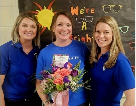 Three women in blue shirts pose next to each other, one holding a bouquet of flowers. Behind them is a wall with a sun and text.