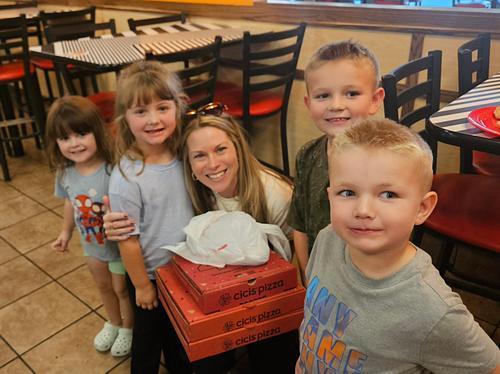 Five children, including a woman, stand in a restaurant. The woman holds boxes of pizza. A tiled floor and tables with red and white checkered tablecloths are visible in the background.