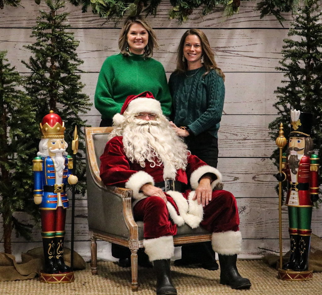 Two women stand behind a man in a Santa suit. He sits in a chair, posing for a photo. Behind them, a backdrop of festive decorations.