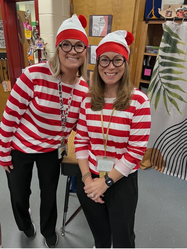 Two women in red and white striped long-sleeve shirts, black pants, and white beanies. They are smiling and standing in a room.