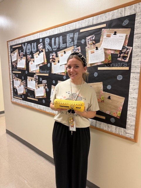 A woman stands in front of a bulletin board displaying a "You Rule" theme. She holds a yellow ruler and wears beige shirt and black skirt