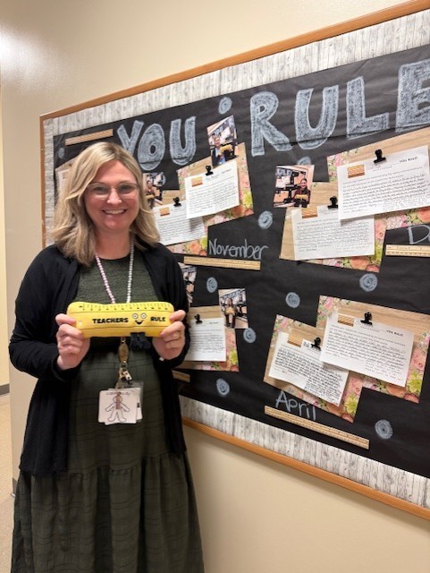 A woman stands in front of a bulletin board displaying a "You Rule" theme. She holds a yellow ruler and wears glasses and a green dress.