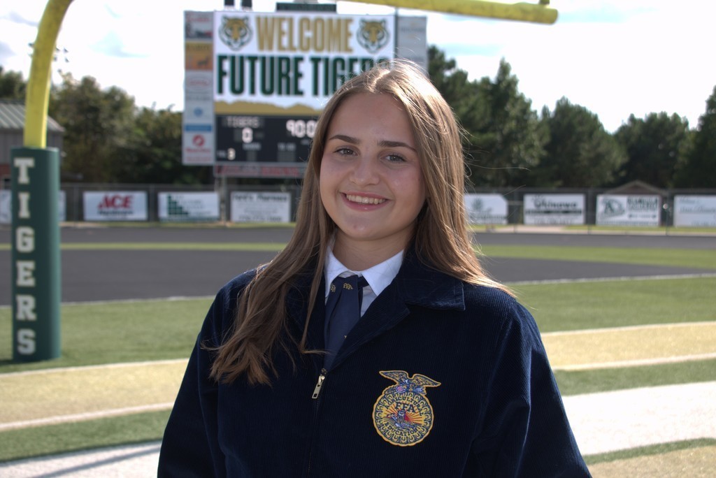 A young woman with long hair and a school uniform stands on a sports field with a large sign.