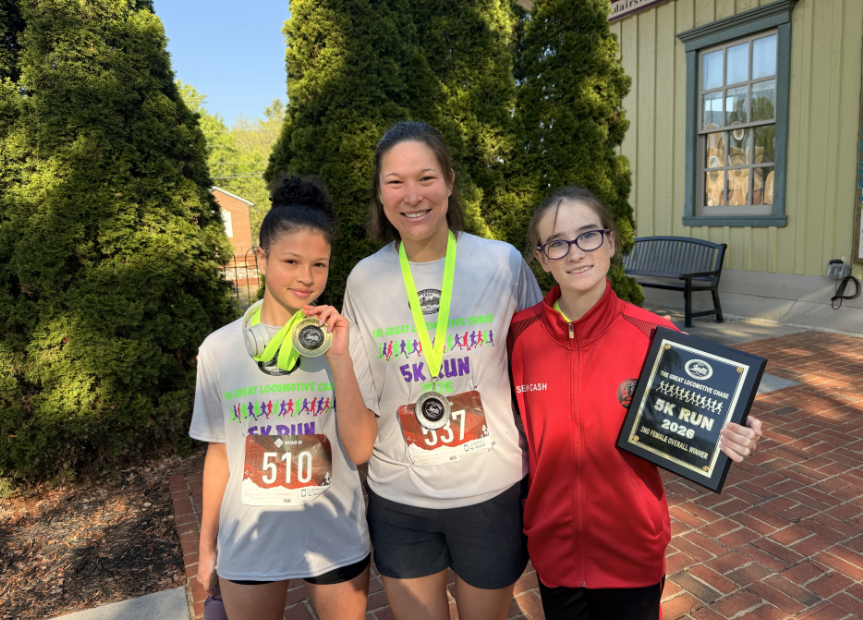 Three individuals stand on a brick sidewalk, smiling for a photo. The girl on the right holds a plaque, while the other two wear medals
