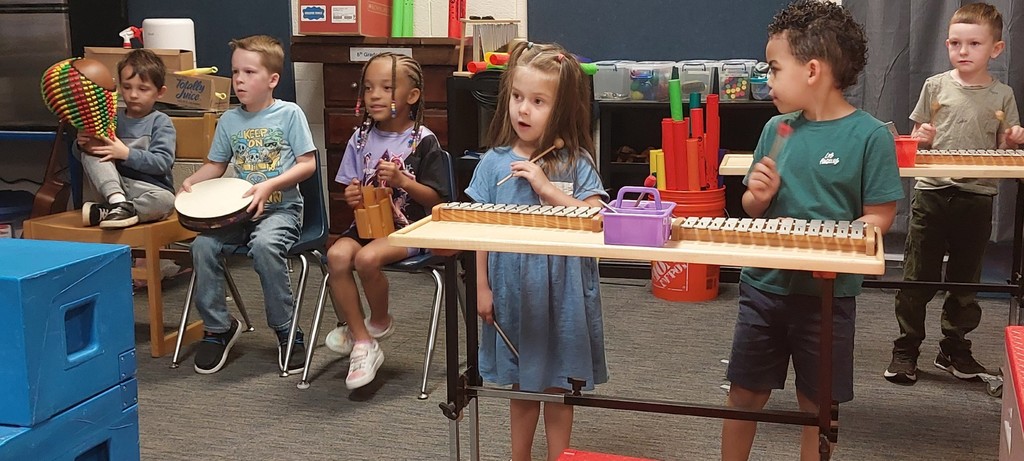 Group of children seated and standing around instruments. Some hold drums and sticks.
