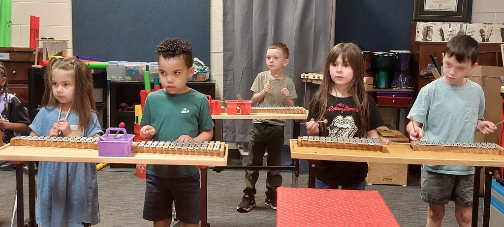 Children playing xylophones in a classroom with a blue wall and curtains in the background.