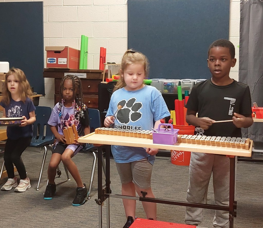 Four children, one standing, play instruments in a classroom. One child plays a xylophone.