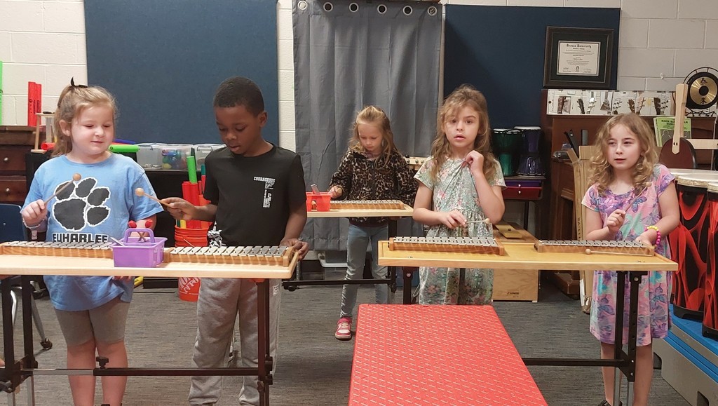 Five young children in a classroom setting, playing musical instruments on a table. Two children are using xylophones.