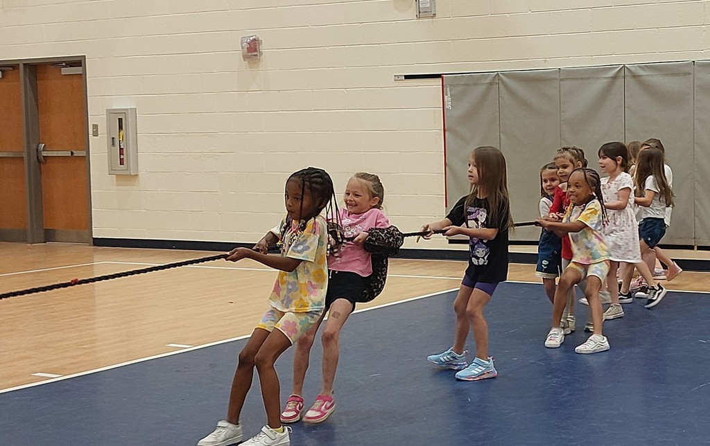 Several children play tug-of-war on a blue mat in an indoor gym, with walls and doors in the background.