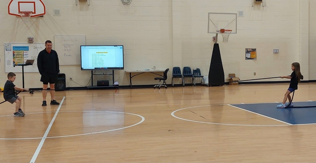Three people on a gym floor, two children pulling on a rope while an adult watches.