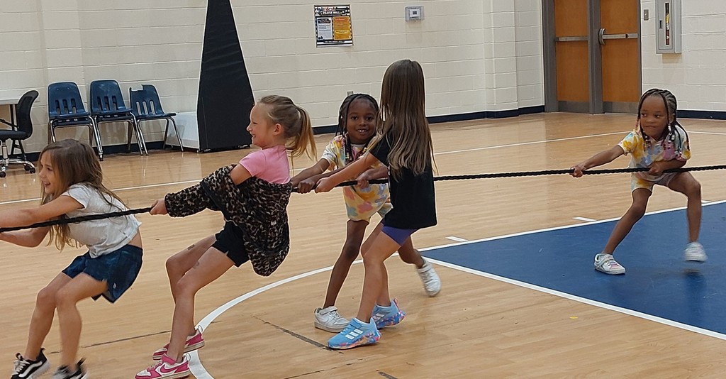 Five young girls in a gym with a wooden floor and blue markings, pulling on a long rope.