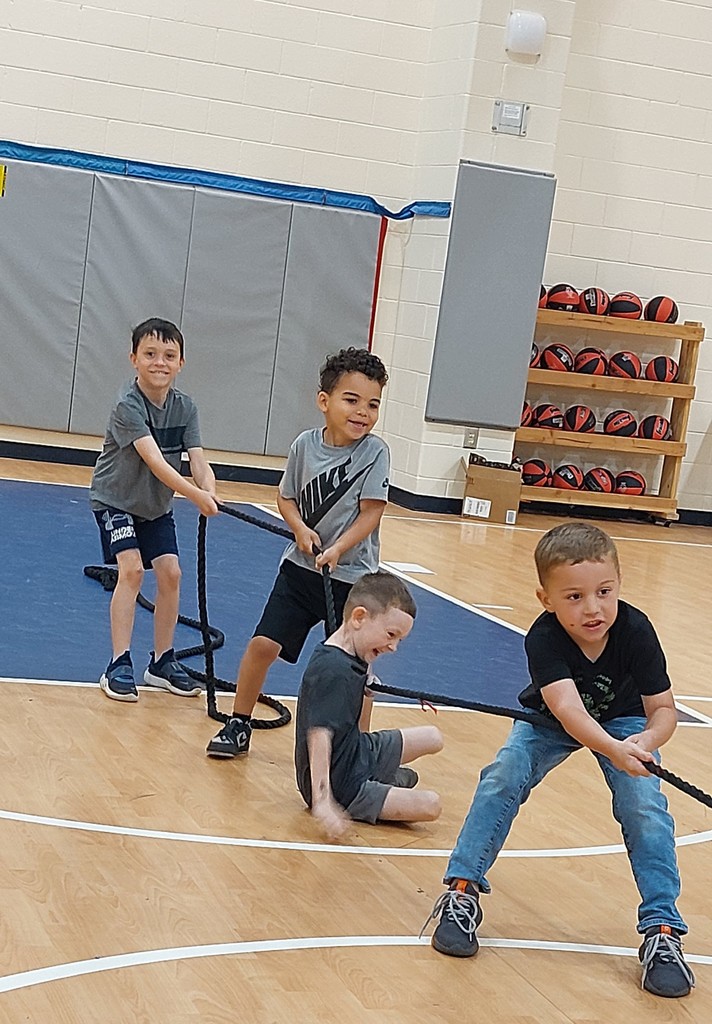 Four young children engage in a tug-of-war game in a gym, wearing casual clothes and sneakers.