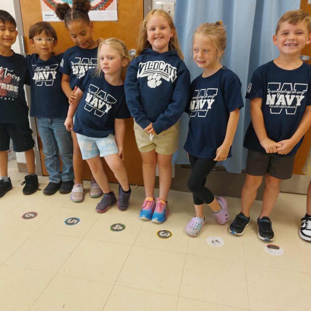 Nine children in navy blue shirts lined up, smiling, in a hallway with blue curtains.