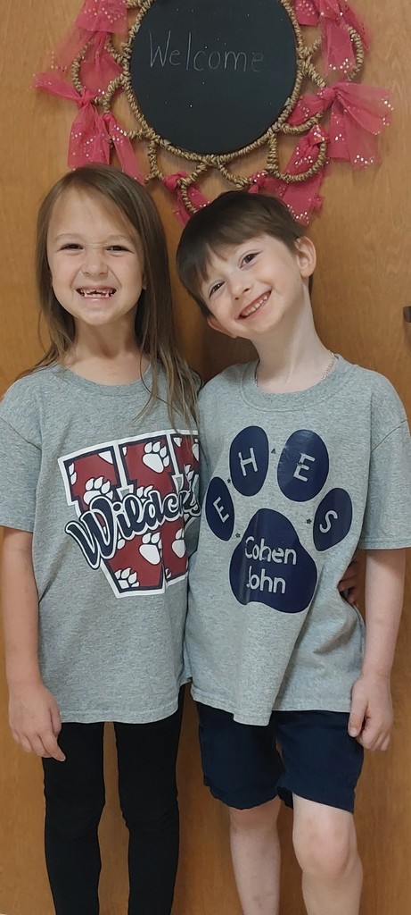 Two children, a girl and a boy, stand together. They wear gray T-shirts with paw prints and school names. Behind them is a wooden door with a decorative sign that reads "Welcome".