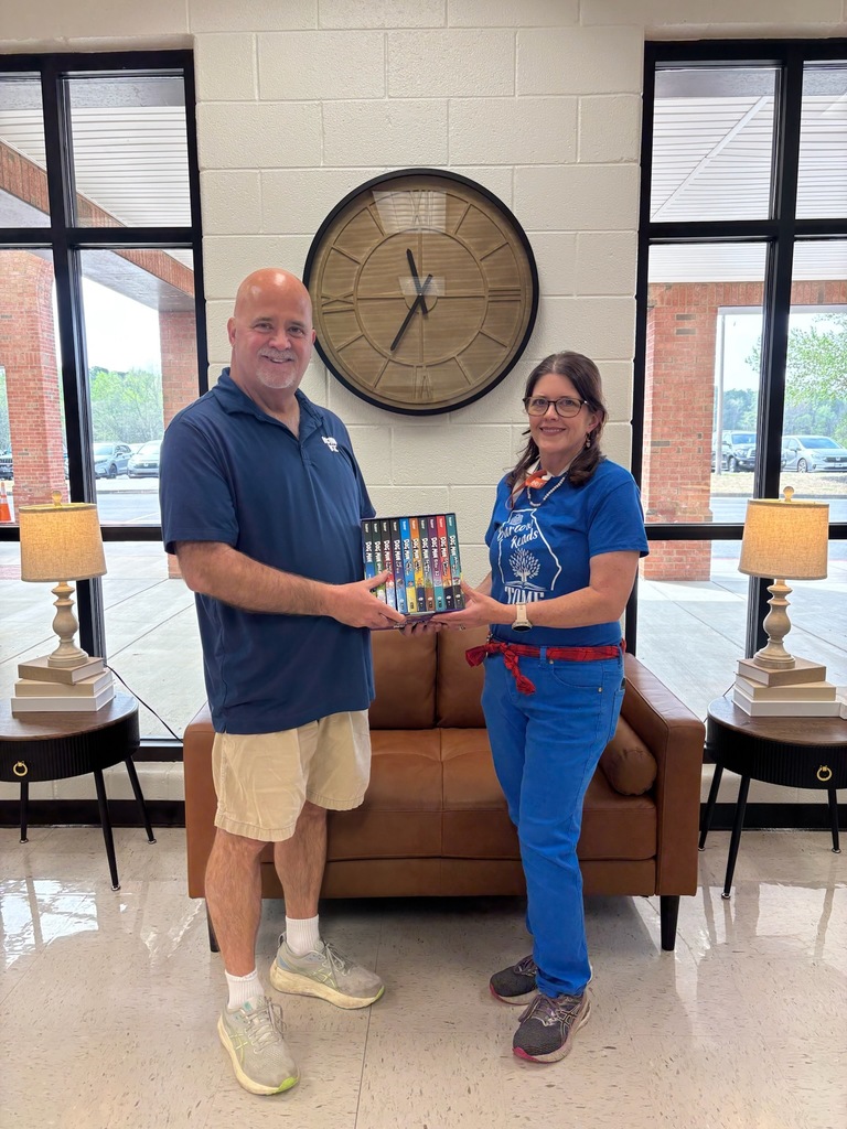 Two adults stand in a school lobby, holding a colorful box of books together in front of a couch and wall clock.