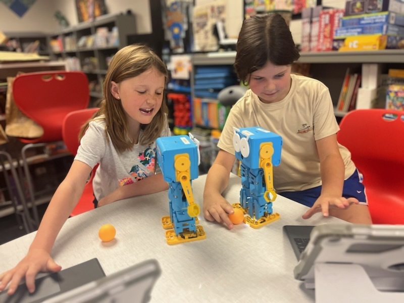 Two children seated at a table, each with a small Marty robot. The robots are facing each other, and one student is holding a ping pong ball