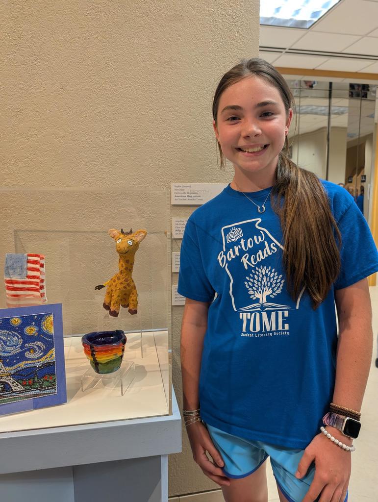 A smiling girl stands next to a display case with a giraffe sculpture, an American flag, and colorful pottery. She has on a blue shirt that says Bartow Reads TOME Reading Society.