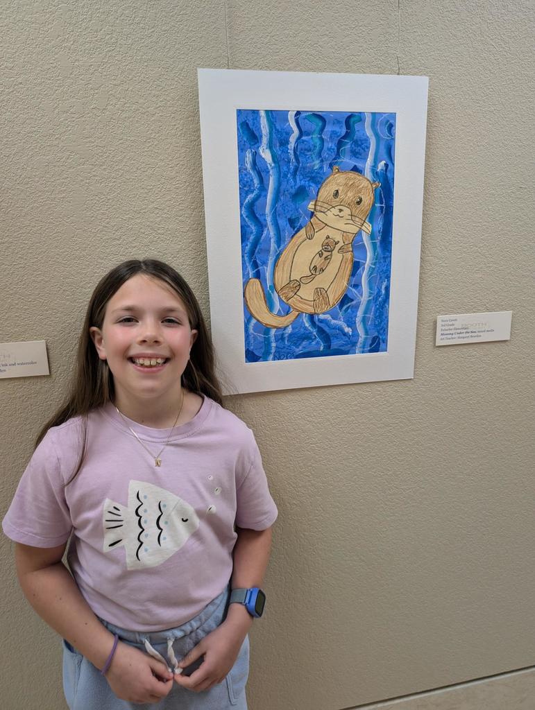 A smiling girl stands next to a framed art piece depicting a sea otter in blue water.