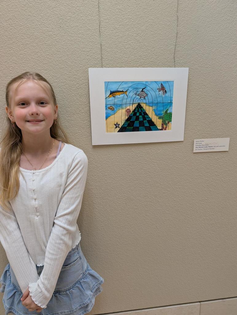 A girl with blonde hair stands by a wall, smiling, next to a framed artwork of a pyramid and sea life.
